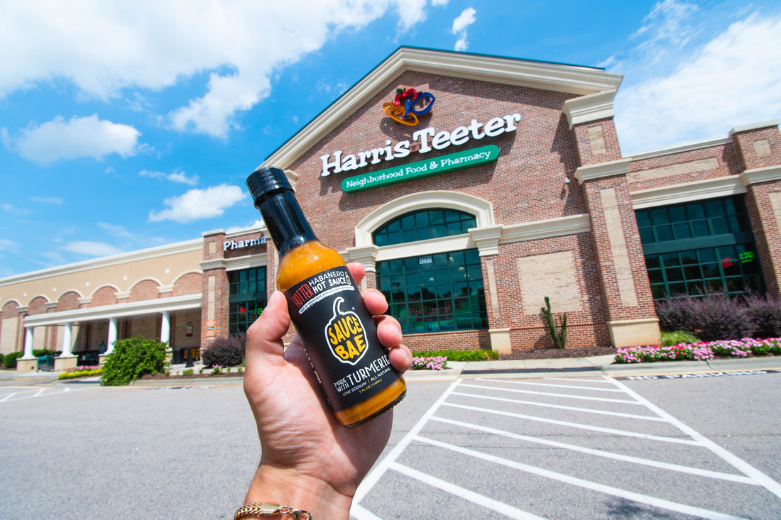 Picture of a hand holding a bottle of Sauce Bae Hotter Habanero Hot Sauce in-front of a Harris Teeter Grocery Store in North Carolina