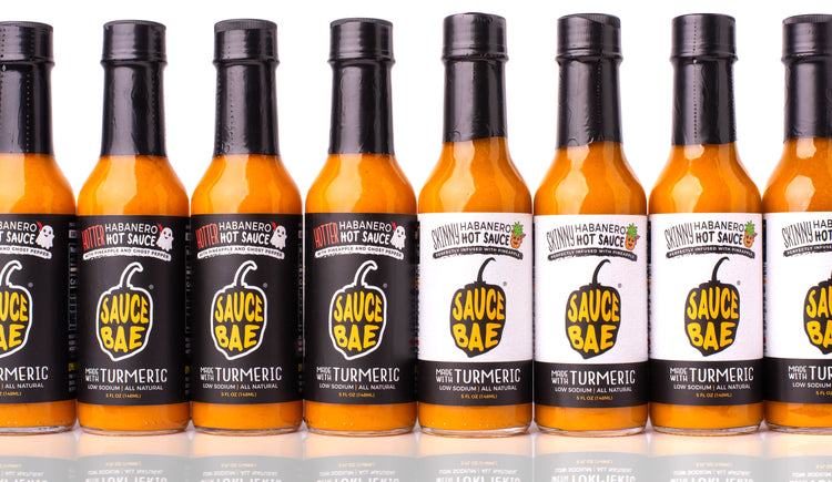 Lineup of Sauce Bae hot sauces featuring alternating bottles of Hotter Habanero with black labels and Skinny Habanero with white labels. Each 5 oz bottle displays the Sauce Bae logo and “Made with Turmeric” tagline. The hot sauces are arranged in a clean row against a white background with subtle reflections below, showcasing the visual contrast between the two flavors.